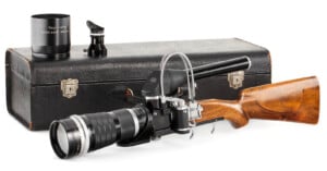 Vintage camera with a telephoto lens attached to a rifle stock for stabilization, alongside additional lenses and a carrying case.