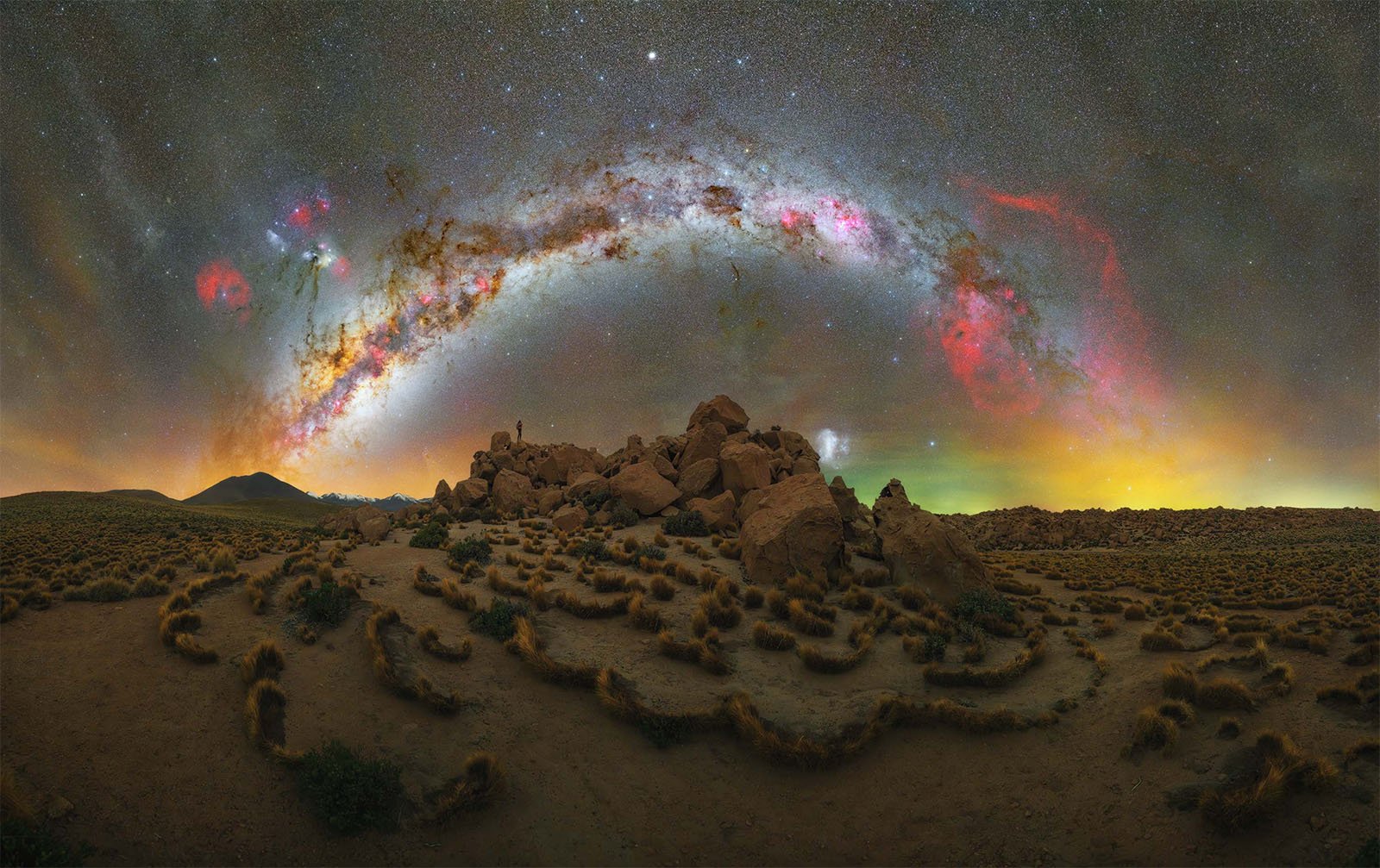  Panoramic view of the night sky with a vivid Milky Way arc stretching across a desert landscape. The ground is dotted with tufts of grass and a rocky formation sits at the center. The sky is illuminated with stars and colorful nebulae, presenting a stunning cosmic scene.
