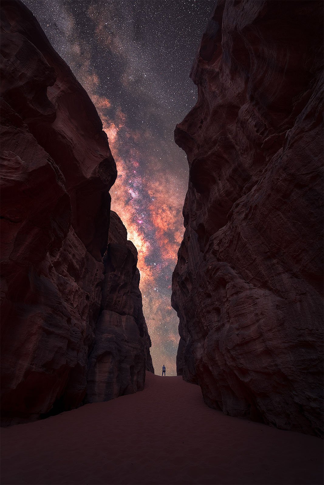  A person stands at the end of a narrow canyon looking up at a vividly colorful, star-filled night sky. The towering rock walls of the canyon contrast with the bright, sprawling Milky Way galaxy above. The scene evokes a sense of awe and wonder.