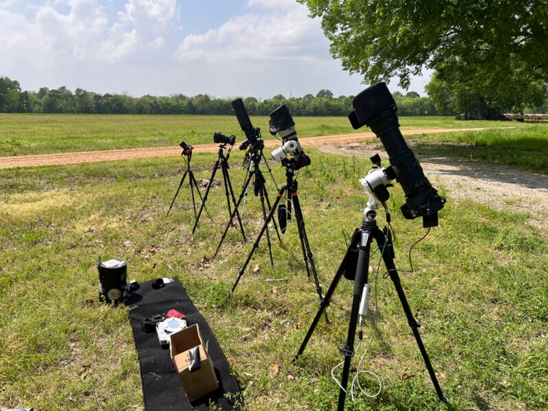 A group of telescopes and cameras on tripods are set up in a grassy field under a partly cloudy sky. Nearby, there is a box and some equipment laid out on a black cloth. Trees and a dirt path are visible in the background.