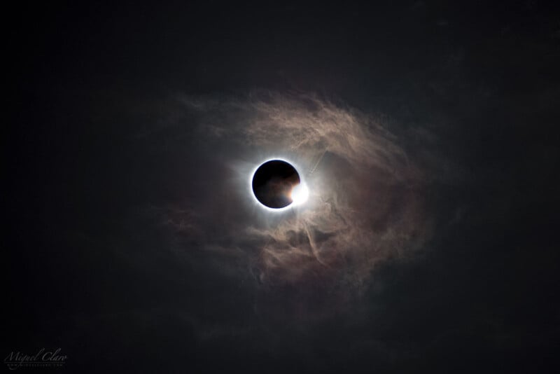 A solar eclipse with the moon partially covering the sun, creating a vivid corona and a bright diamond ring effect. Surrounding clouds add a dramatic touch, enhancing the contrast against the dark sky.