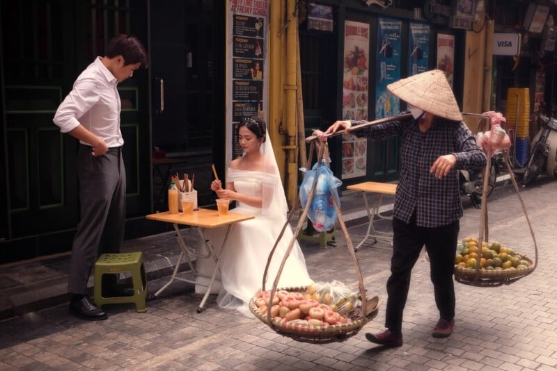 A bride in a white dress sits at a small outdoor table beside a man adjusting his shirt on a busy street. A vendor carrying a traditional shoulder pole with baskets of fruit walks by, blending modern and traditional elements in an urban setting.