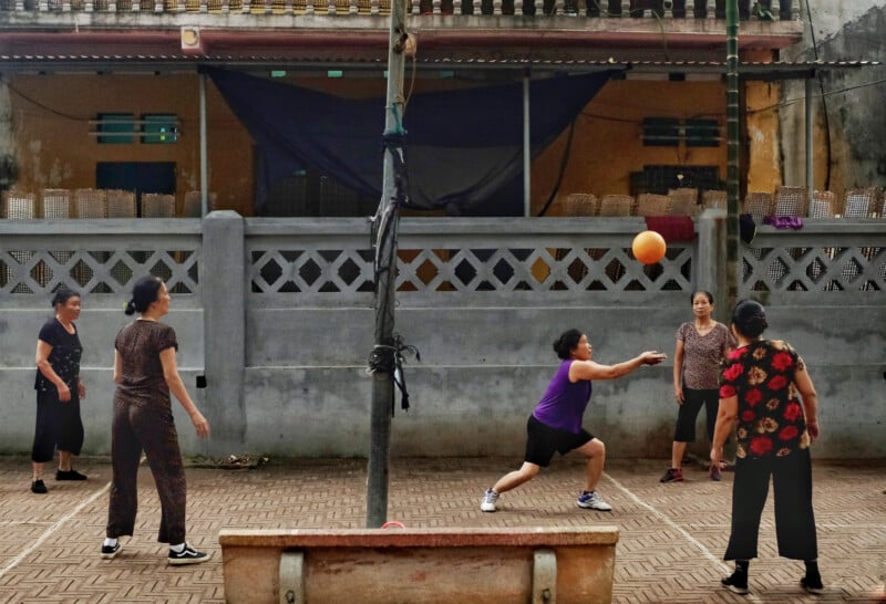 A group of women are playing volleyball in an outdoor courtyard bordered by a low wall. One woman is poised to hit the ball, while others observe. They are dressed casually, and buildings with balconies and a shaded area are visible in the background.