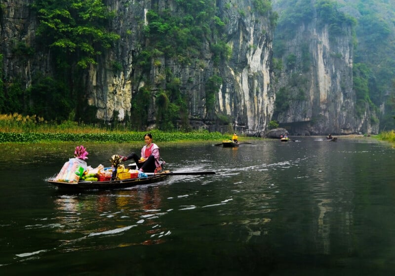 A person navigates a small boat filled with goods along a tranquil river surrounded by towering limestone cliffs and lush greenery. Other boats can be seen in the distance, with a misty atmosphere adding to the scenic beauty of the landscape.