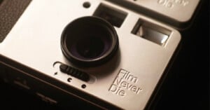 Close-up shot of a retro-style camera. The camera has a circular lens, a small flash, and a textured grip on the left. It features a selector switch beneath the lens, and the words "Film Never Die" engraved on the right side. The design suggests a vintage aesthetic.