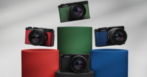 A display of four Lumix cameras in different colors (green, red, blue, and black) mounted on cylindrical pedestals of corresponding colors. The background is gradient grey, and the cameras are presented at varying heights.