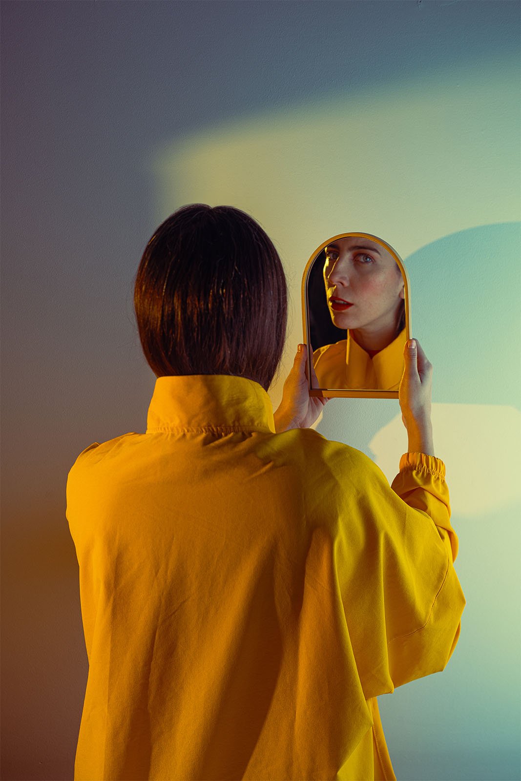 A person with shoulder-length dark hair wearing a yellow shirt stands facing away and holds a small circular mirror, reflecting their face. The background is a gradient of blue, green, and yellow. The scene is dramatically lit, casting vivid shadows.