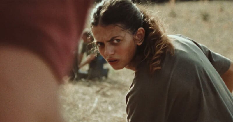 A woman with her hair tied back looks intensely over her shoulder. She is outdoors in a dry, open area. Other people are visible in the blurred background, sitting or crouching on the ground. The mood appears tense or serious.