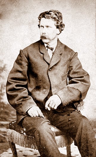 A black-and-white vintage photograph of a man seated on a wooden chair. He has short, wavy hair and a mustache and is wearing a formal suit jacket over a shirt with a bow tie. His hands rest on his legs, and he is gazing slightly to his left.