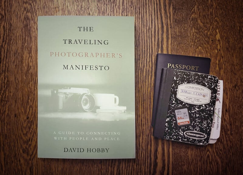 A book titled "The Traveling Photographer's Manifesto" by David Hobby is placed on a wooden surface. Next to it, there is a blue passport and a small composition notebook labeled "Composition Hand Legend" with some travel tickets attached.