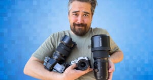 A person with a beard and mustache holds three cameras against a blue, pixelated background. They have a playful and slightly bewildered expression, wearing a light-colored t-shirt. One camera has a long lens, and the other two have shorter lenses.