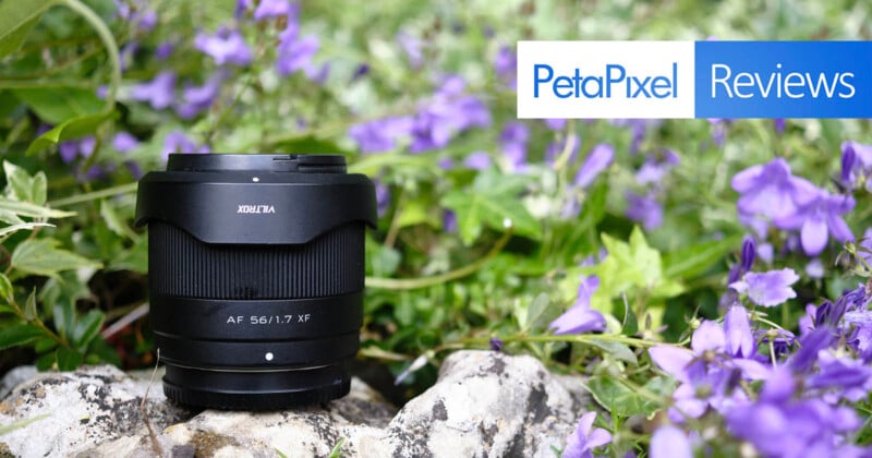 A black camera lens sits on a rock surrounded by vibrant purple flowers and green foliage. In the top right corner of the image, the PetaPixel logo is displayed alongside the word "Reviews" on a blue background.