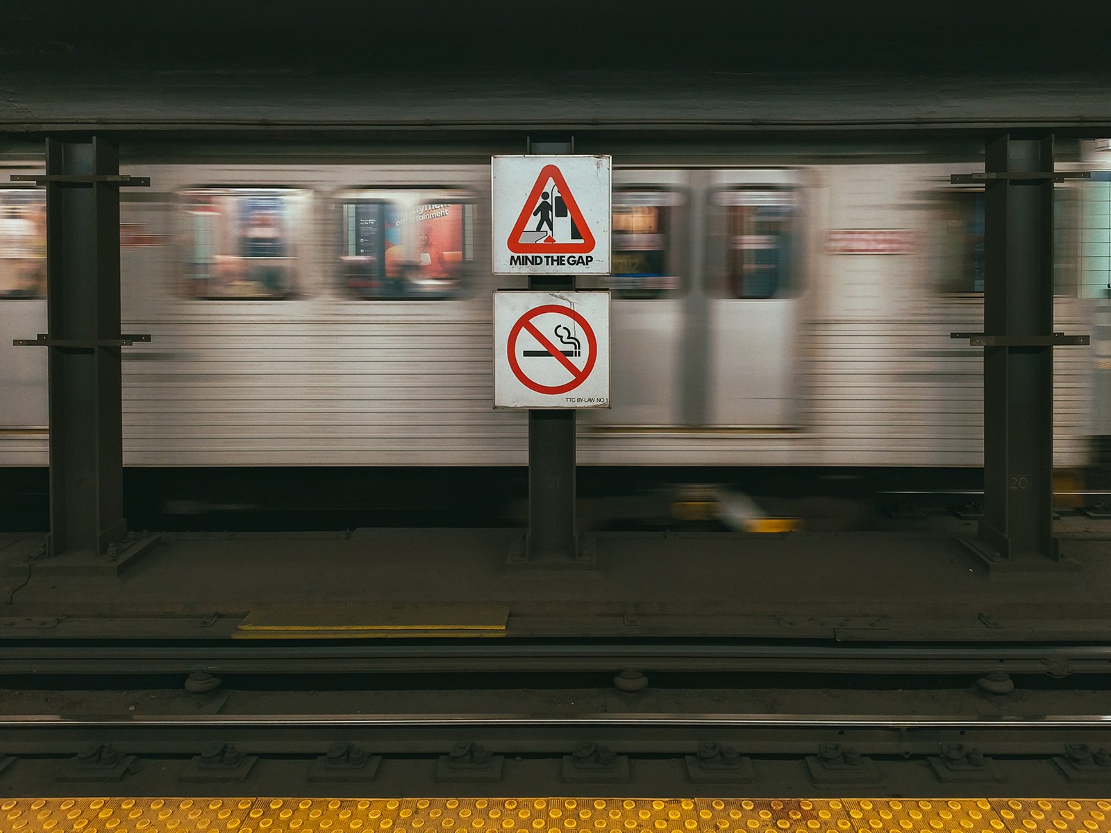 A motion-blurred subway train passing behind a platform sign with warnings "mind the gap" and "no smoking" at a station. yellow safety line visible on platform edge.