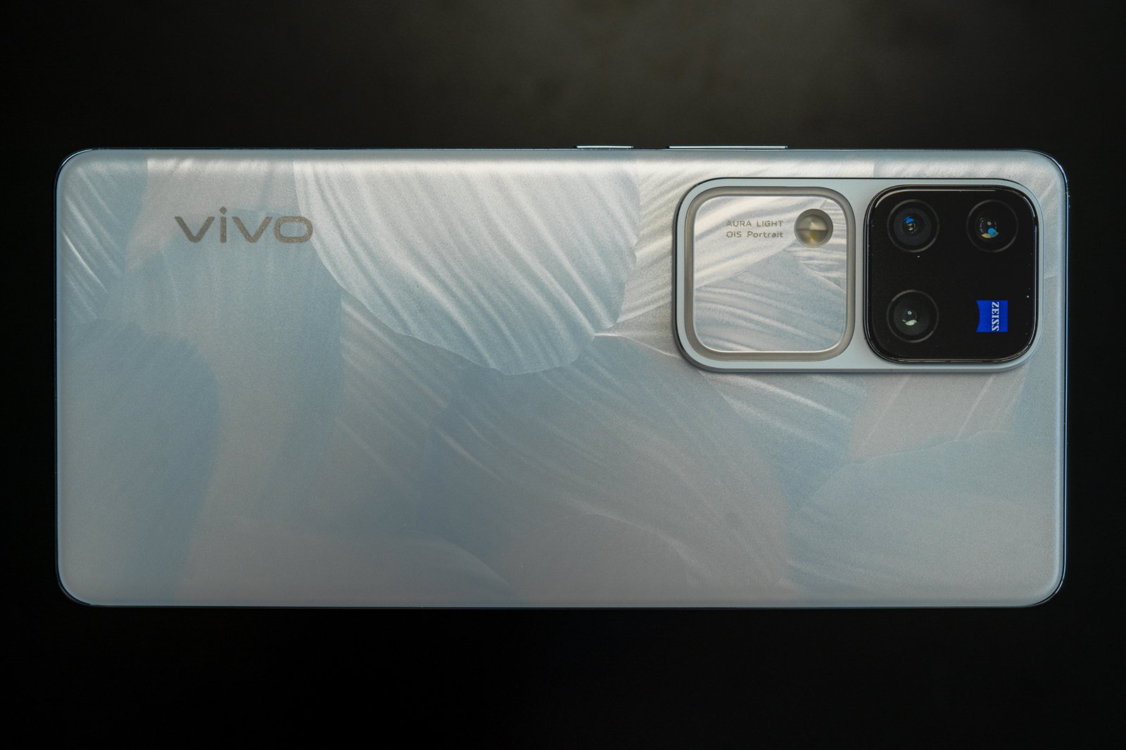 A vivo smartphone with a reflective silver finish lies on a dark surface. it features a large, square camera module with multiple lenses and a blue zeiss logo.