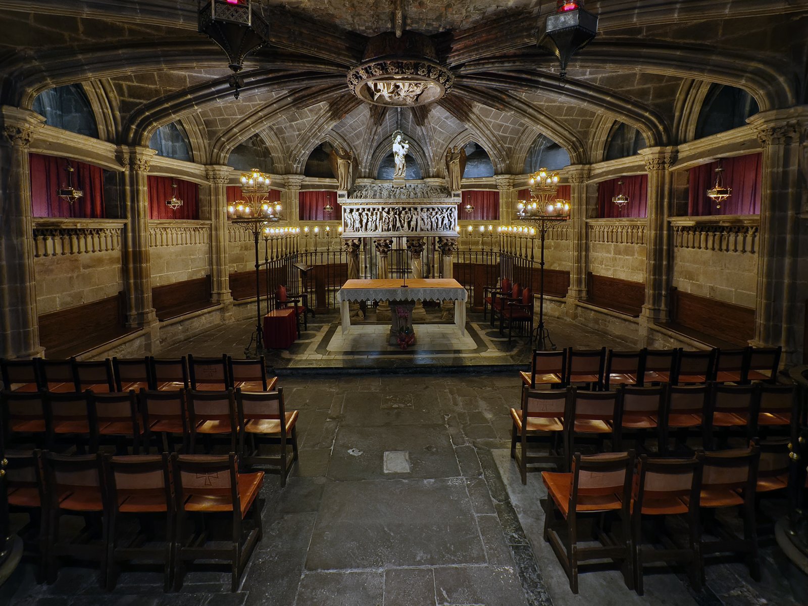 Interior of a gothic chapel with vaulted ceilings, wooden pews, and an ornate altar featuring a statue of a figure in a blue robe. decorative stonework and a chandelier enhance the historical ambiance.