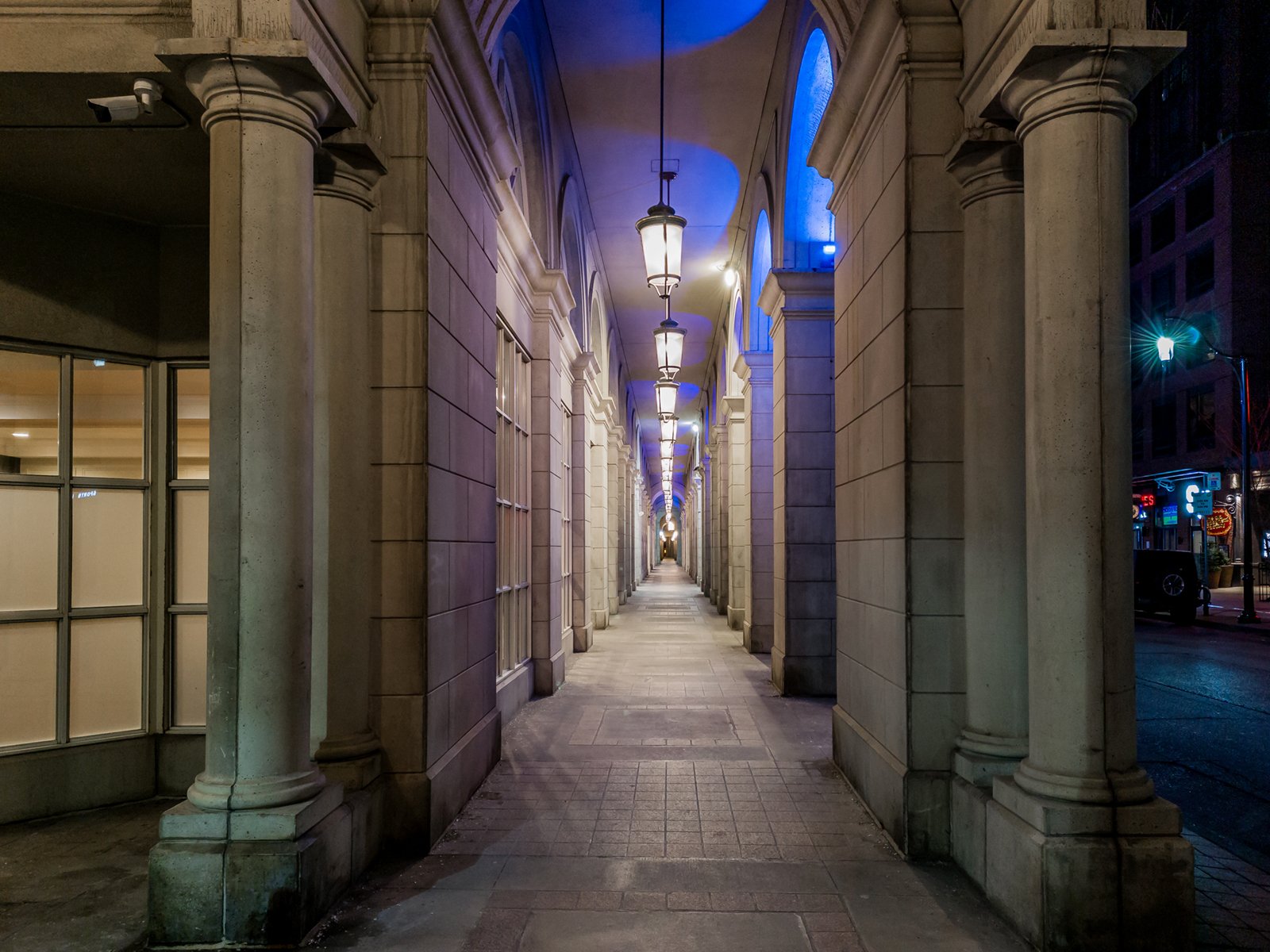 A narrow passage between classical stone arches, illuminated by hanging lanterns at dusk, leading to a vivid blue-lit archway in the distance. the path is deserted and creates a serene urban scene.