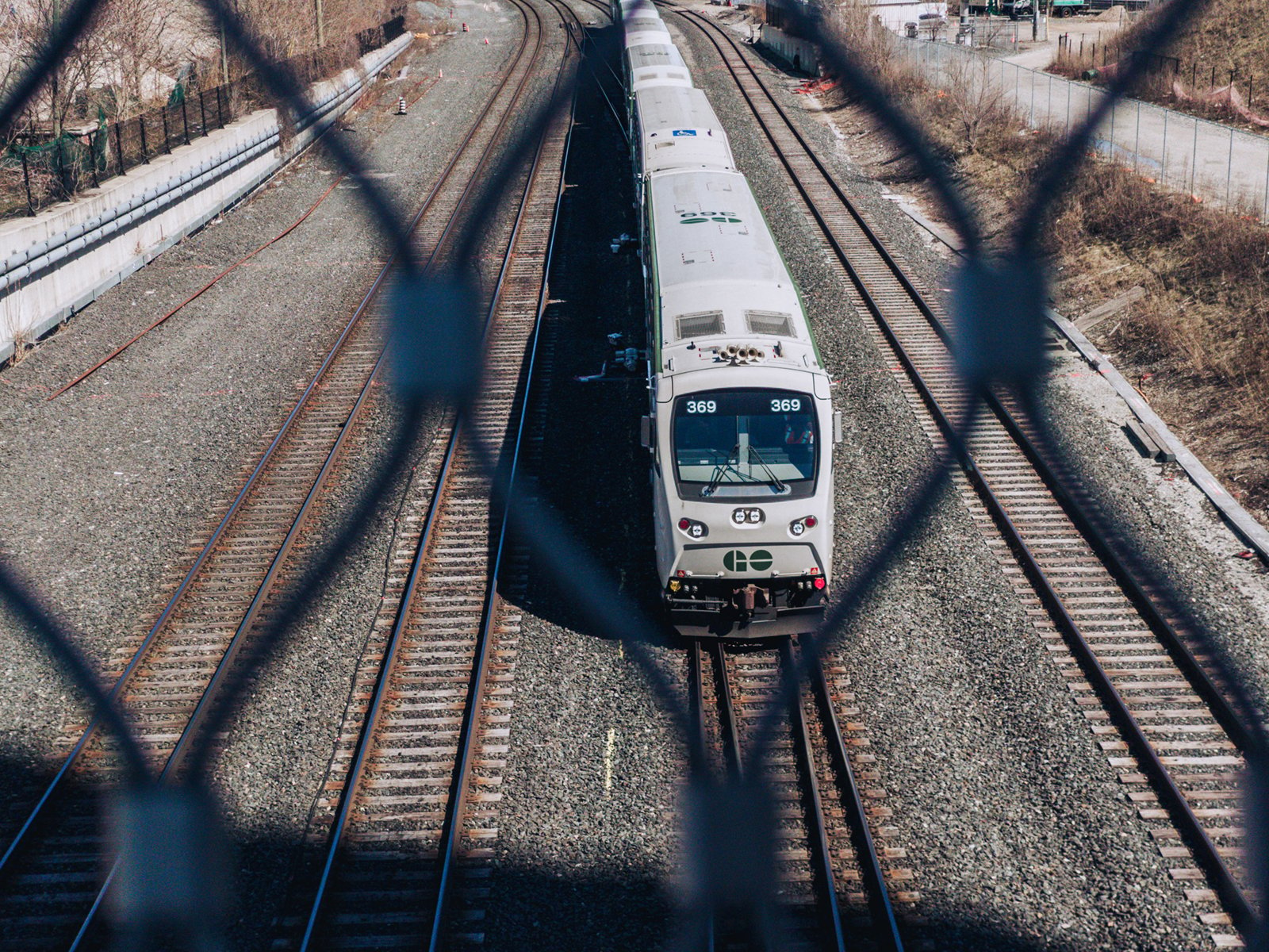A commuter train seen through a chain-link fence, traveling on multiple tracks, captured from an overhead perspective. the train, predominantly white, moves away from the viewer.