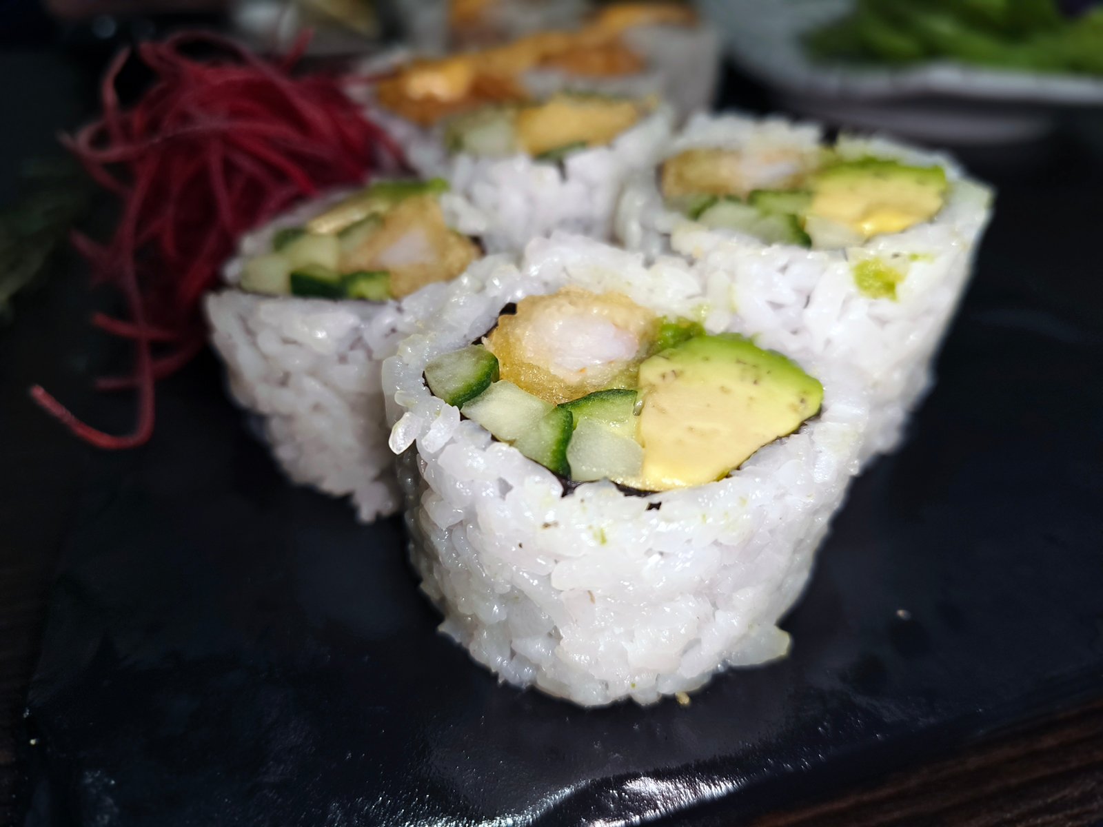 Close-up image of sushi rolls with avocado and cucumber on a black plate, garnished with red threads on the side.