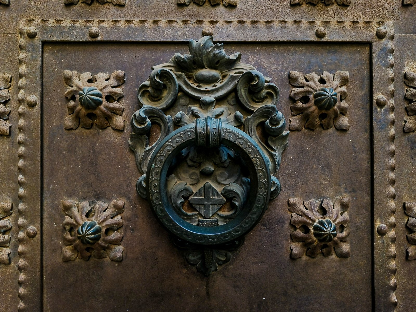 Ancient ornate metal door knocker featuring a lion's head surrounded by decorative floral patterns on a weathered iron door.