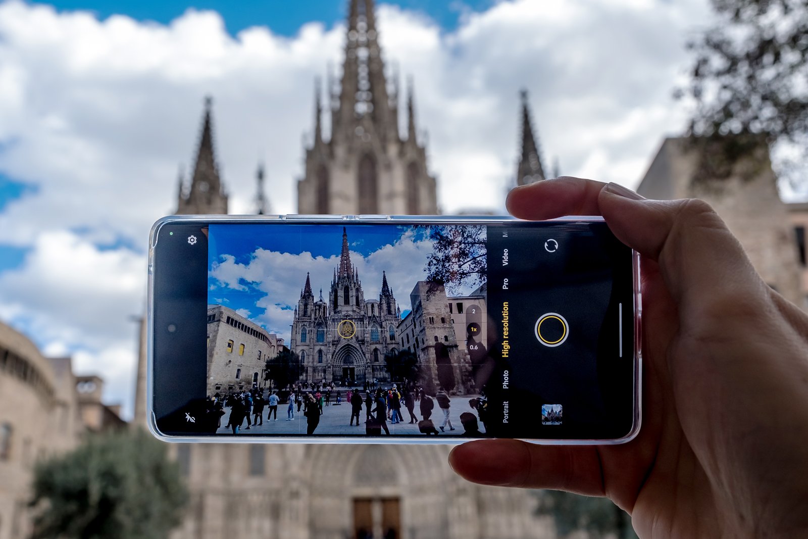 A hand holding a smartphone capturing a photo of a bustling square in front of a grand gothic cathedral under a cloudy sky. the phone screen displays the cathedral image clearly.