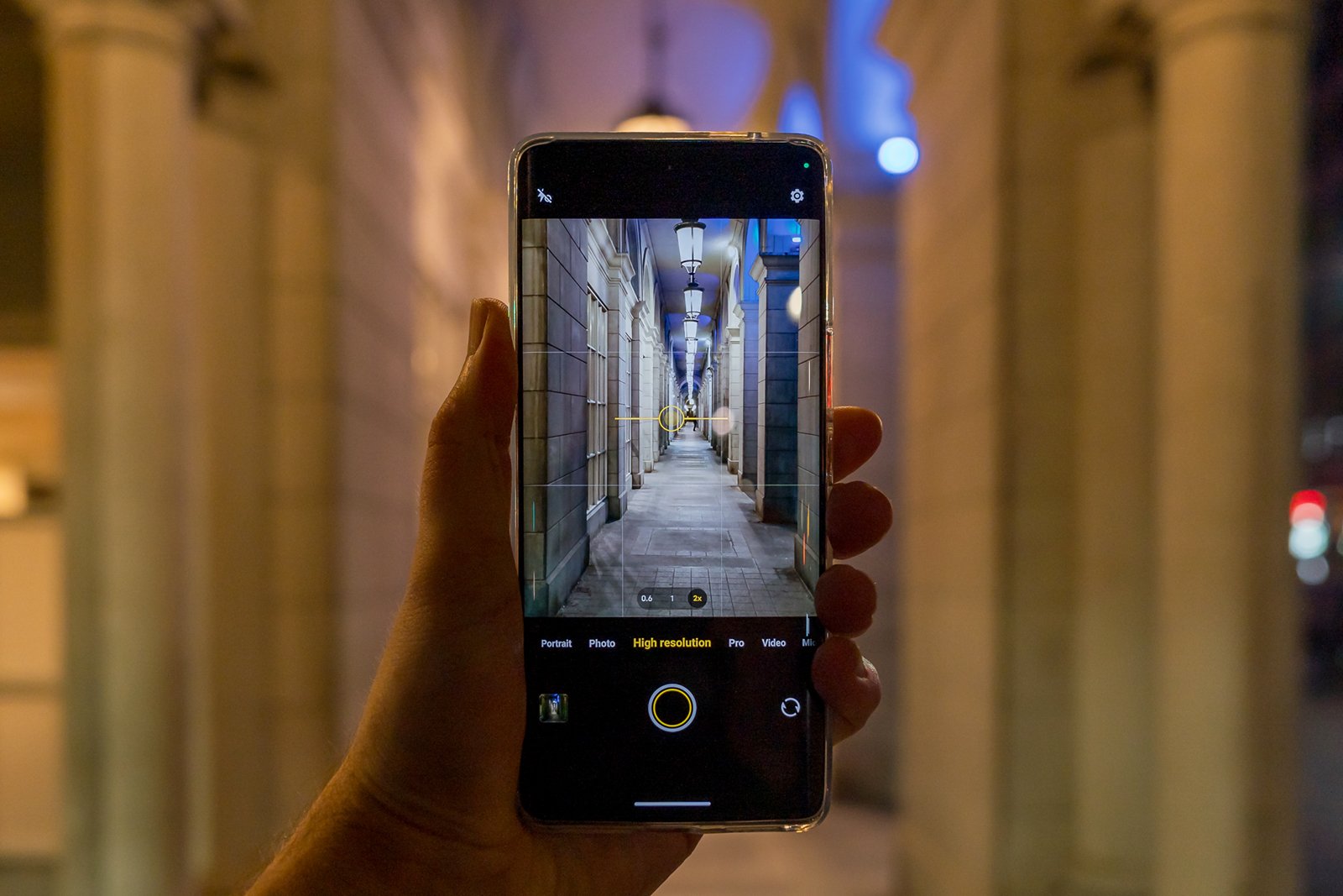A person's hand holding a smartphone, displaying a photograph of a dimly lit corridor with arches on its screen. the real corridor aligns with the image on the phone, creating a framed view.