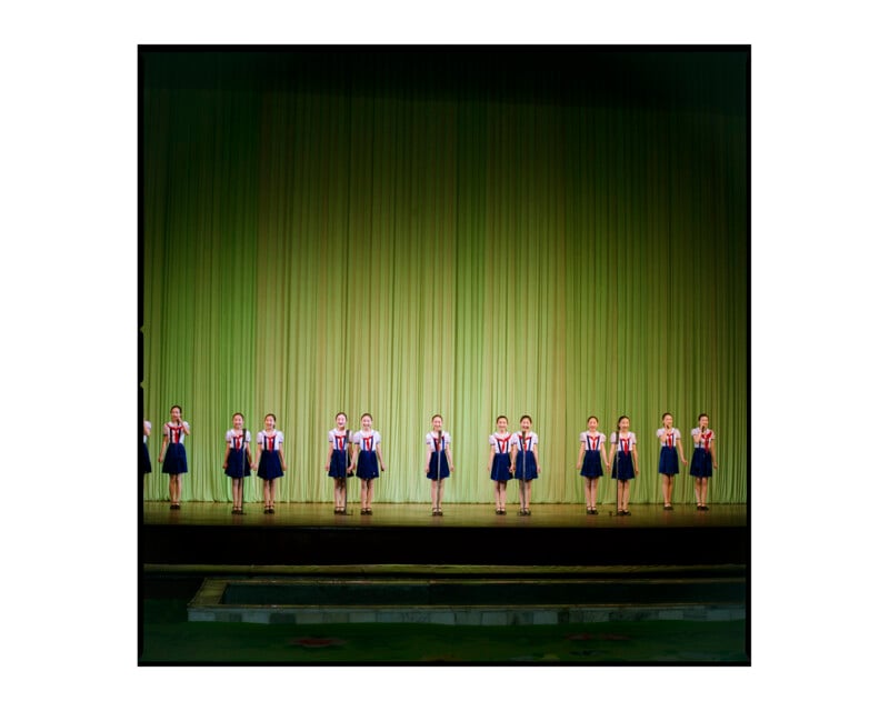 A line of young girls wearing matching blue and white uniforms standing on stage, with a green curtain in the background, during a performance or presentation.