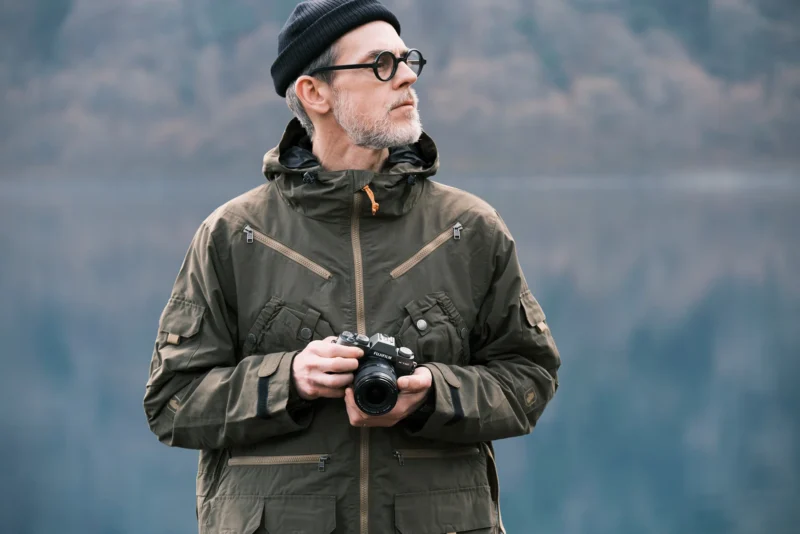 A person wearing a dark beanie, glasses, and a green jacket holds a camera while looking to the side. The background is an out-of-focus natural setting with what appears to be a lake and trees.