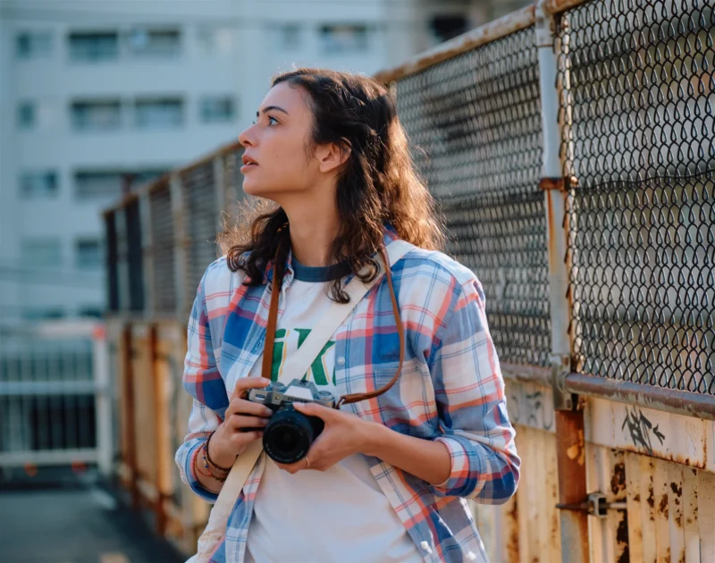 A person with long curly hair, wearing a plaid shirt over a white t-shirt, stands outdoors holding a camera. They appear thoughtful as they look to the side. The background shows a rusty fence and blurred buildings.