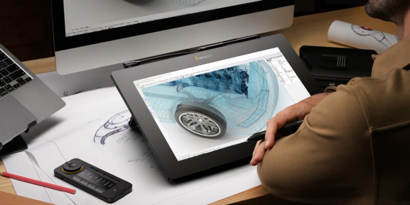 A person is working on a digital drawing of a car wheel displayed on a graphics tablet, with design blueprints scattered around the table and a computer in the background.