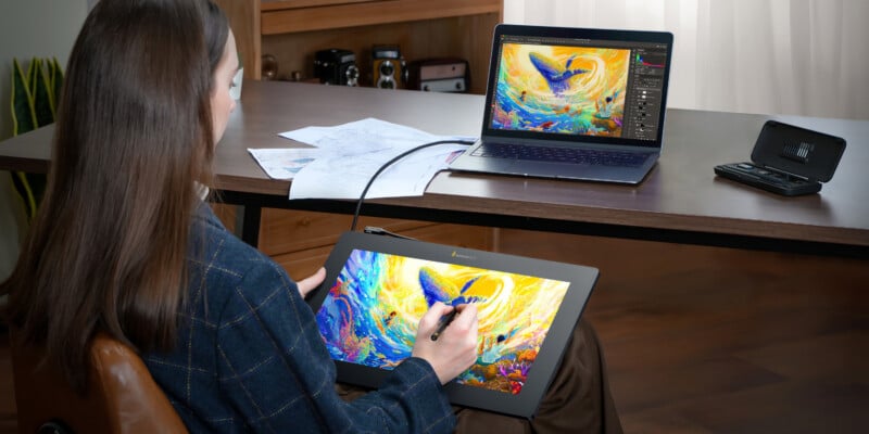 A woman using a stylus on a tablet to edit a colorful abstract artwork, which is also displayed on her laptop screen, in a home office setting.