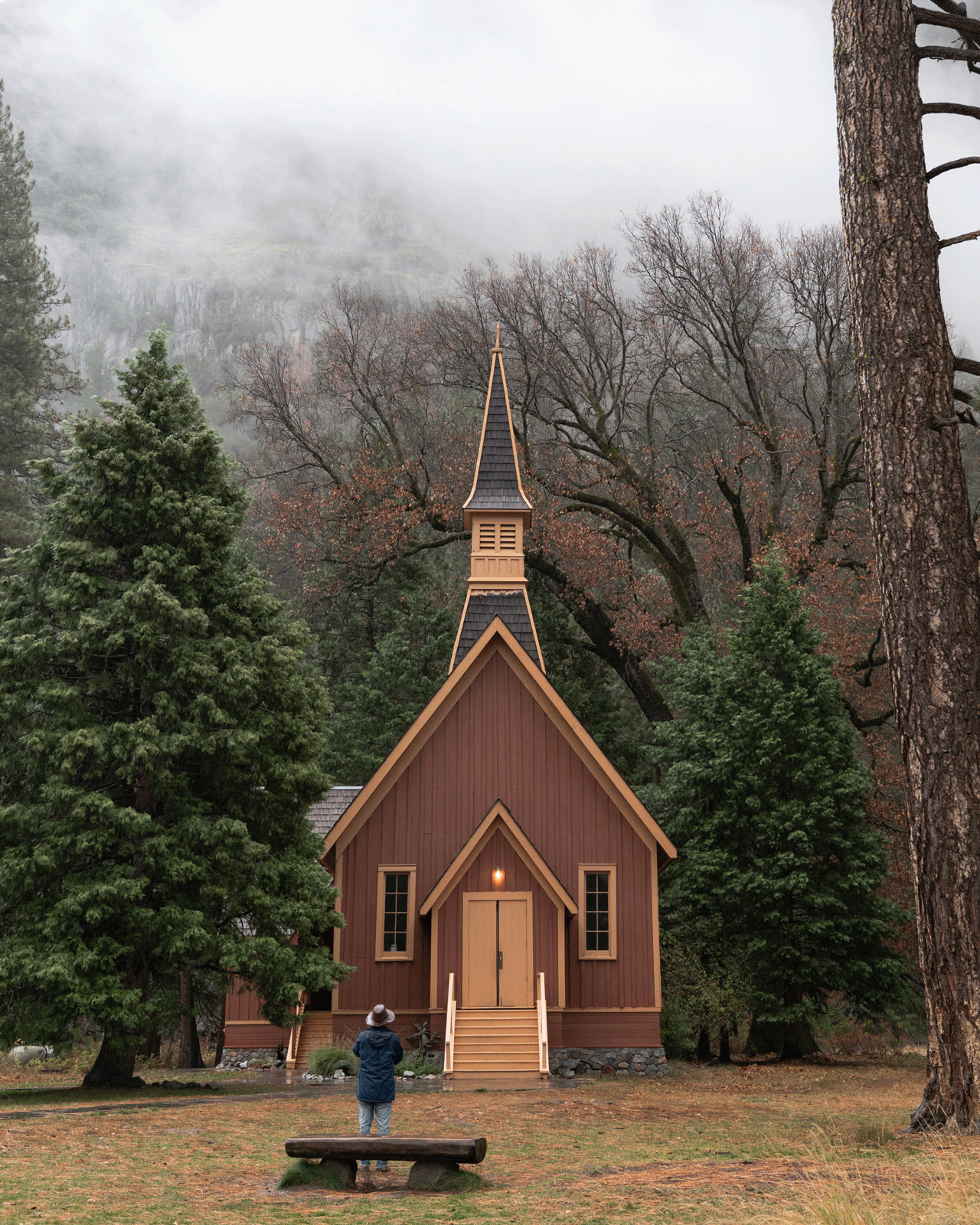 A person stands facing a small, rustic wooden church with a tall steeple, surrounded by trees in a forest. The scene is misty, with low clouds in the background, creating a serene and peaceful atmosphere.