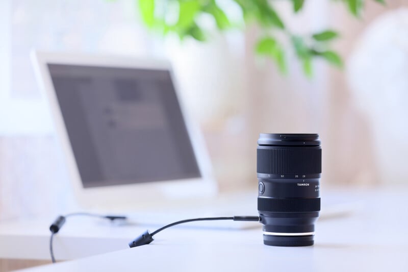 A camera lens is connected to a laptop via a USB cable on a white desk. The laptop in the background has a blurred screen, and there is green foliage out of focus in the background.