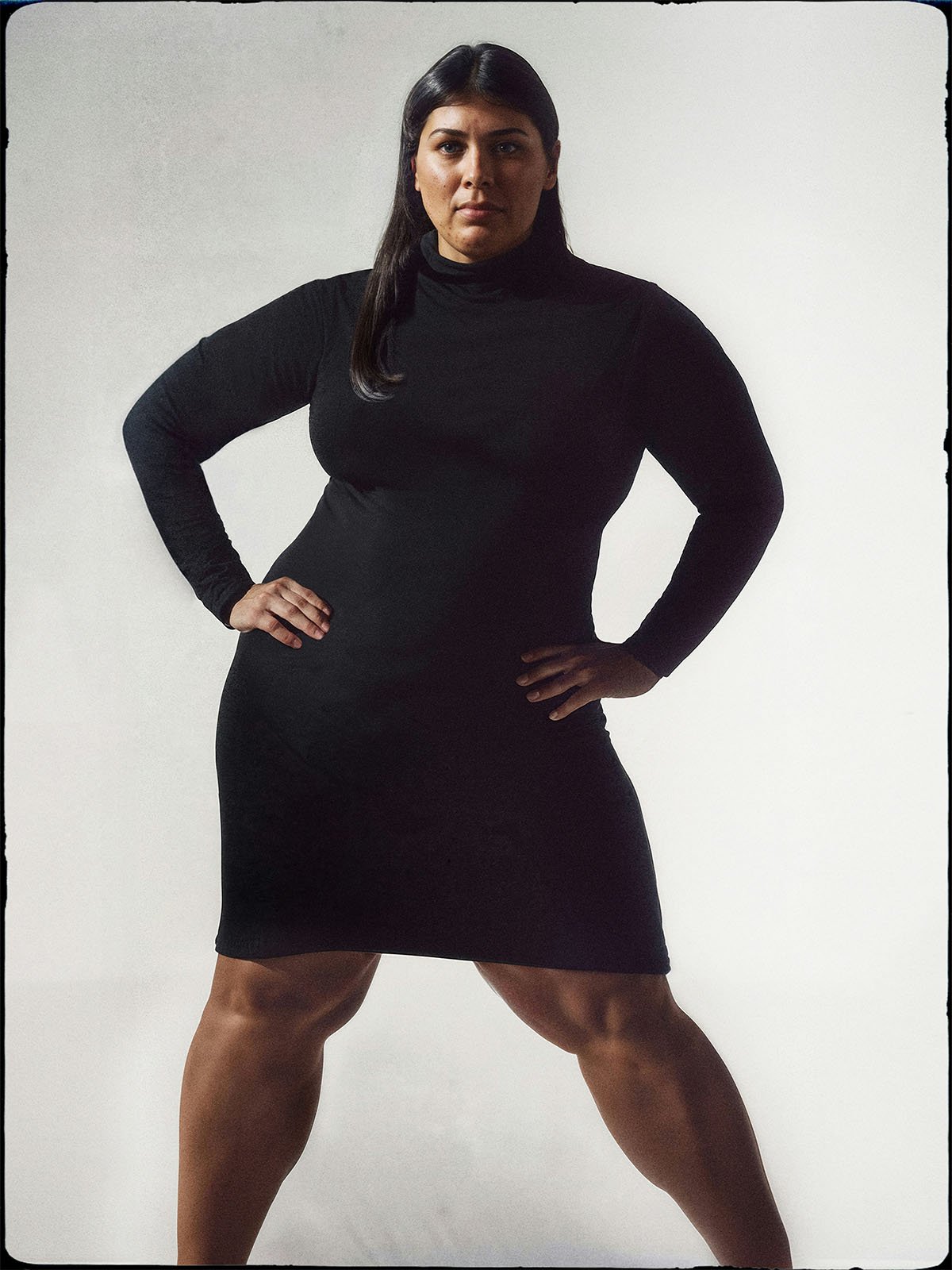 A confident woman in a black turtleneck dress stands with hands on her hips against a light grey background. her expression is assertive and poised.