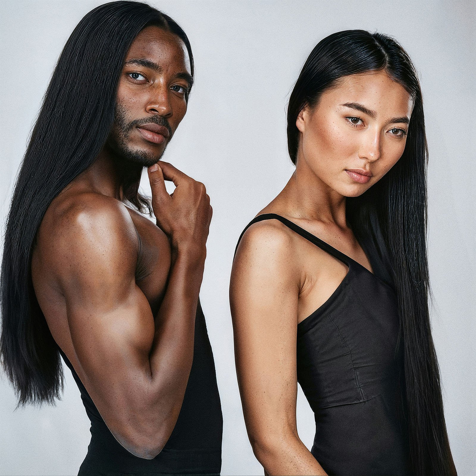 Two models with long straight hair posing back-to-back, one black man shirtless and one asian woman in a black dress, against a grey background.