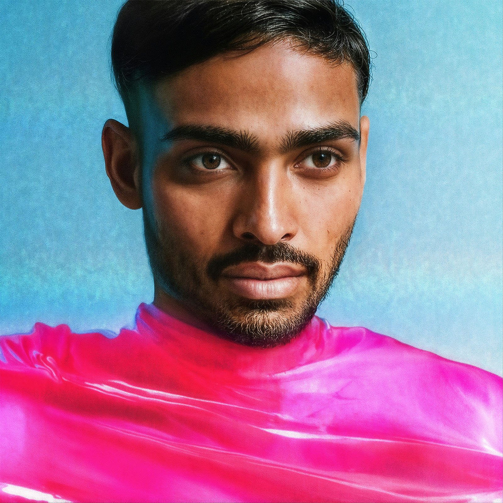 Portrait of a man with facial hair, looking directly at the camera, wearing a bright pink garment. the background is a soft blue texture.