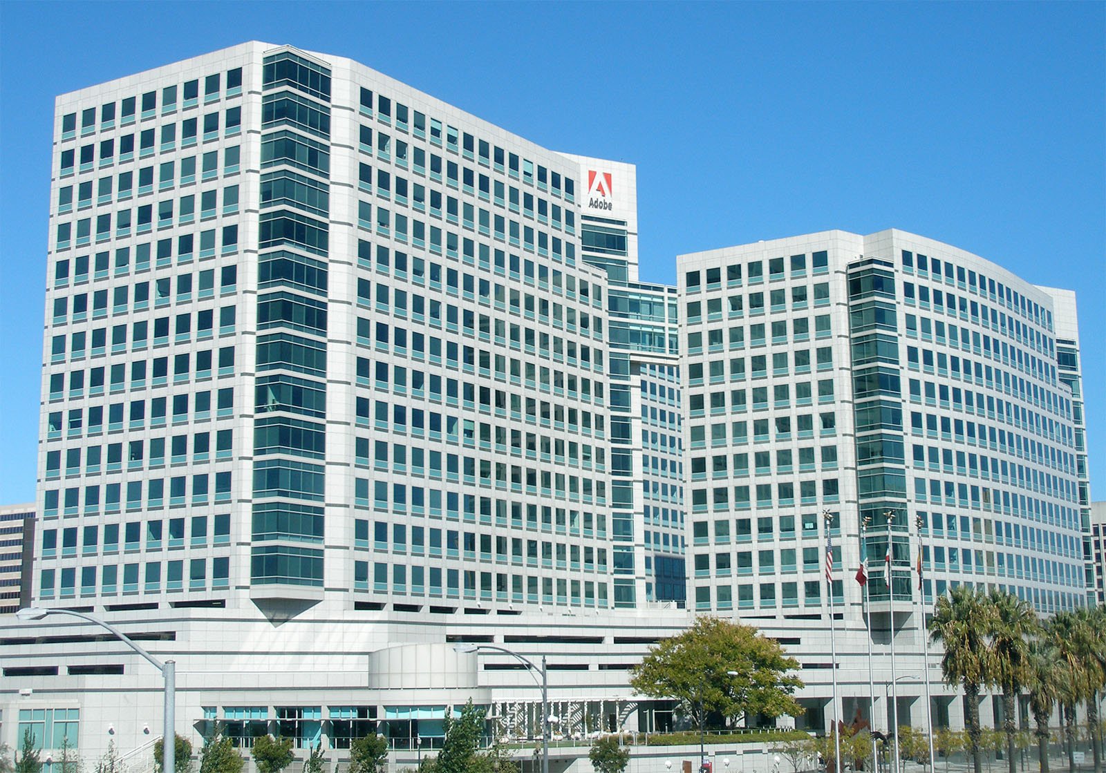A view of the Adobe Systems headquarters, featuring two modern high-rise office buildings with numerous windows. The buildings are white with grey accents and are connected by a glass bridge. Flags are displayed near the entrance, and the sky is clear and blue.