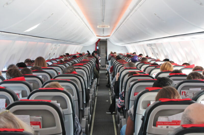 Interior of an airplane showing passengers seated in rows, focusing down the aisle towards the back of the cabin. each seatback has a safety instructions card.