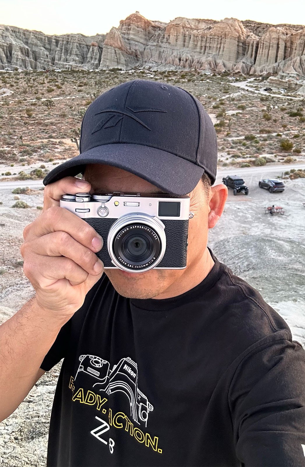 A person wearing a black cap and a black t-shirt with the text "READY. ACTION." is holding a camera up to their face while standing outdoors in a desert-like area with rocky formations and parked vehicles in the background.