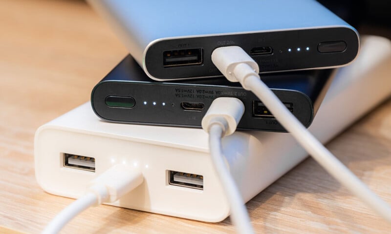 Three stacked portable power banks in varying sizes and colors connected to charging cables on a wooden surface.