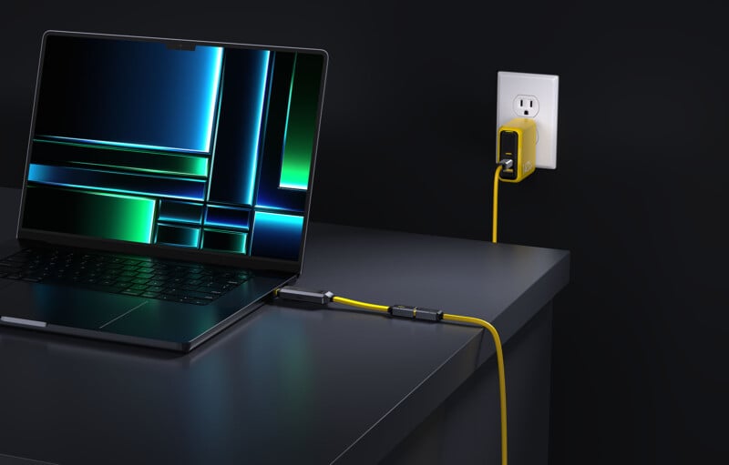 A laptop with a vibrant, abstract screen is placed on a dark desk. It is connected to a power outlet on the wall through a yellow braided charging cable. The sleek, modern setup contrasts with the black background.
