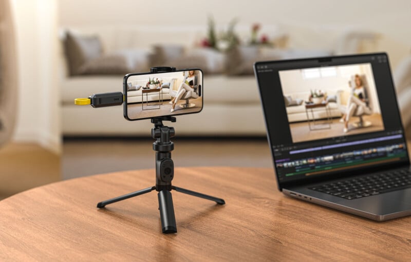 A smartphone mounted on a small tripod on a wooden table captures a video of a person sitting on a couch. The phone is connected to a laptop displaying video editing software, showing the same scene on its screen. The background shows a blurred living room.