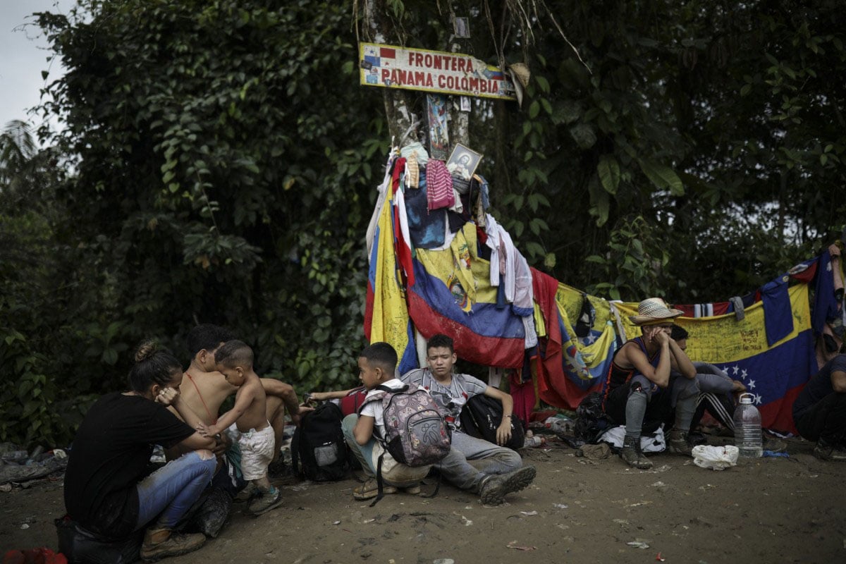 A group of people rest and converse under a sign that reads "frontera panamá colombia," surrounded by various flags and personal belongings in a natural setting.