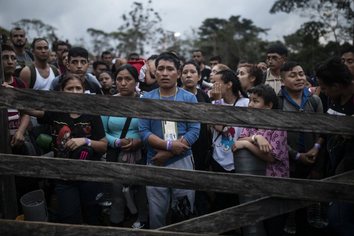 A diverse group of people, some holding children, stand behind a wooden barrier with serious expressions, possibly waiting for assistance or at a checkpoint, in a dimly lit, outdoor setting.