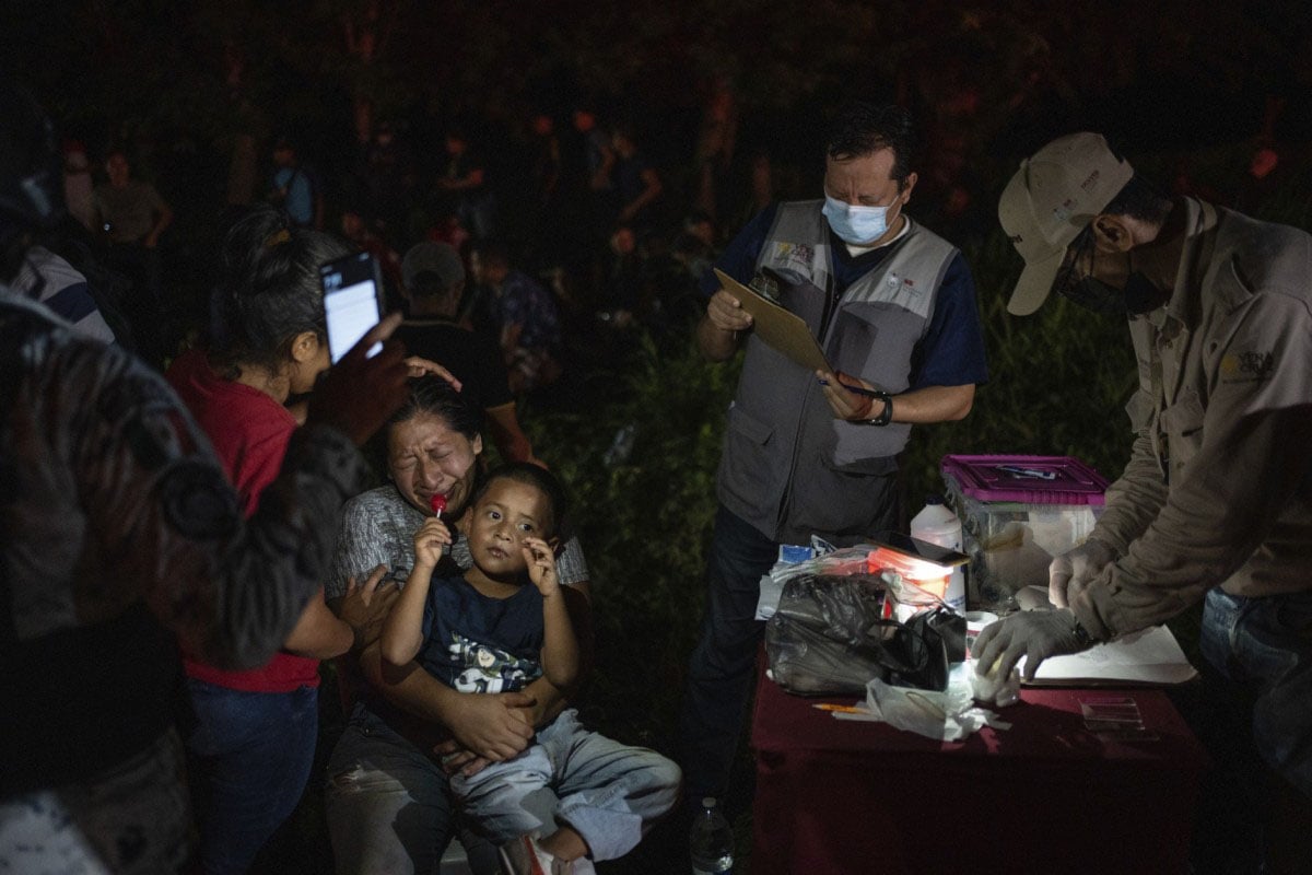 A night scene where medical staff in masks attend to a crying toddler held by a distressed woman. other individuals surround them in a dimly lit outdoor setting.
