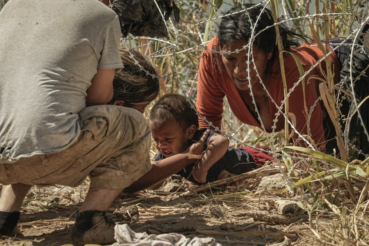 A child crawls on the ground under branches, guided by a man and a woman, who appear focused and concerned, in a natural, rugged outdoor setting.