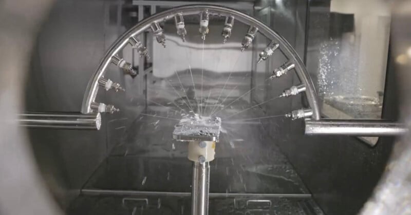 A CNC (Computer Numerical Control) machine operates inside an enclosed workspace, showing multiple nozzles on a curved metal arm spraying coolant on a piece being precisely milled. The surrounding area is wet with coolant splashes.