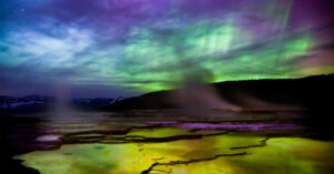 Vivid aurora borealis illuminates the night sky over a mysterious landscape with glowing yellow thermal springs in the foreground, surrounded by dark mountain silhouettes.