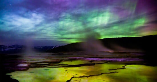 Vivid aurora borealis illuminates the night sky over a mysterious landscape with glowing yellow thermal springs in the foreground, surrounded by dark mountain silhouettes.