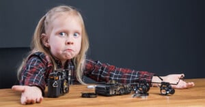 A young girl with long blonde hair in a plaid shirt sits at a table with her arms out in confusion, surrounded by disassembled camera parts. She looks wide-eyed and puzzled, as if unsure how to put the camera back together.
