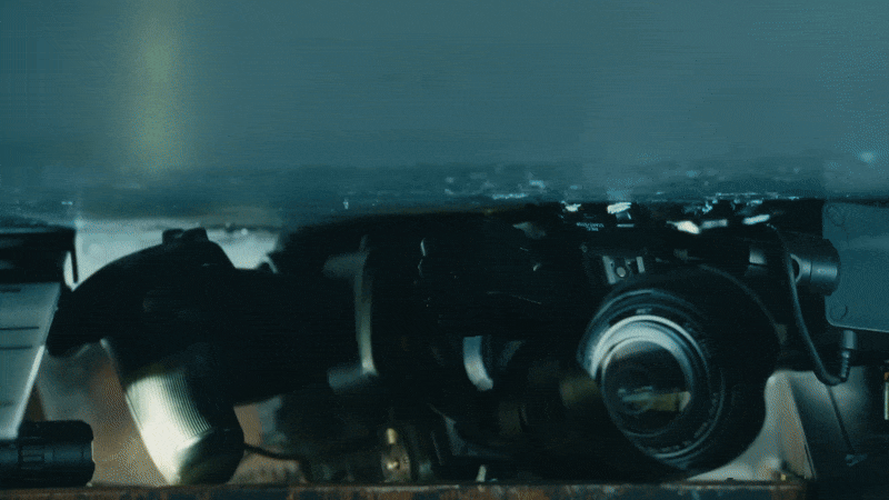 Old camera equipment submerged in shallow, moving water, including cameras and lenses in a worn and possibly discarded condition, highlighted by a soft overhead light.